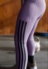 Leggings lilás claros com três riscas verticais pretas de cada lado, feitos de um material elástico, apresentando um ajuste justo. O fundo está desfocado.