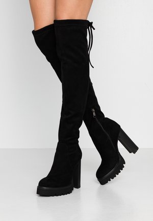 Jambes portant des bottes montantes en daim noir au-dessus du genou avec des talons et des semelles compensées épaisses, fermées par des fermetures éclair latérales, debout sur un sol blanc.