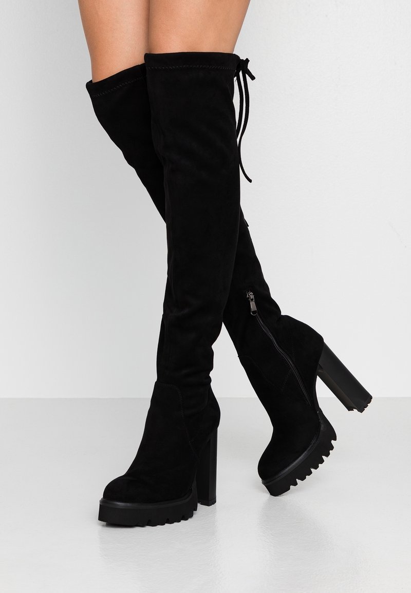 Jambes portant des bottes montantes en daim noir au-dessus du genou avec des talons et des semelles compensées épaisses, fermées par des fermetures éclair latérales, debout sur un sol blanc.