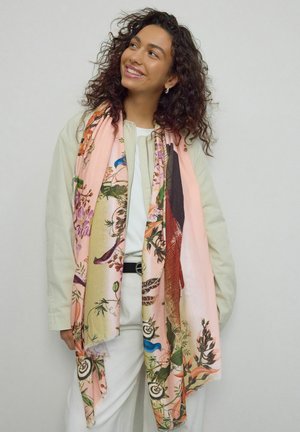 Lächelnde Frau mit lockigem Haar trägt eine leichte Jacke und Hose, ergänzt durch einen großen Schal mit Blumen- und Vogelmustern.