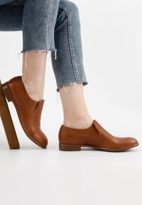 Bruine leren instappers met een ronde neus, lage blokhak en gebogen uitsnijdingen. Draagt met versleten denim jeans.