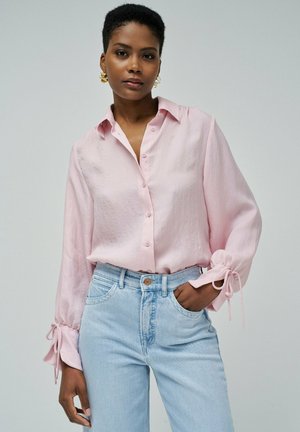Camisa botón de color rosa claro con mangas largas y detalles de lazo en las muñecas, combinada con jeans de tiro alto en color azul claro. Textura de tela suave.