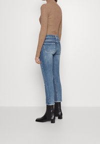 Suéter de cuello alto marrón texturizado combinado con jeans de tiro medio azules y botas negras hasta el tobillo. Cuenta con bolsillos y una silueta ajustada.