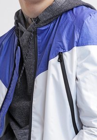 Homme portant une veste zippée bleu et blanc sur un sweat à capuche gris avec des cordons noirs et un tatouage visible au cou.