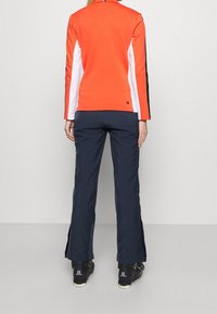 Maglia in pile rossa con pannelli laterali bianchi e neri, abbinata a pantaloni blu navy. Presenta un logo rotondo sul retro e calzature nere.