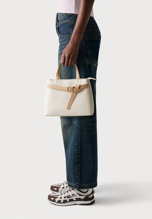 White leather handbag with tan accents, rectangular shape, short handles, and a belt detail. Worn with wide-leg jeans and patterned sneakers.