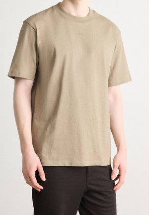 Osoba ubrana w jednolity, beżowy t-shirt z krótkim rękawem z małym wytłoczonym logo oraz ciemne spodnie, widoczna od szyi do bioder na białym tle.