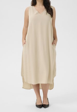 Woman wearing a sleeveless beige midi dress with pockets and black pointed-toe heels, standing against a plain background.