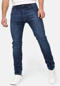 Jeans de denim azul oscuro con un corte ajustado, que presentan un ligero desgaste y un diseño clásico de cinco bolsillos. Combinados con zapatillas blancas.
