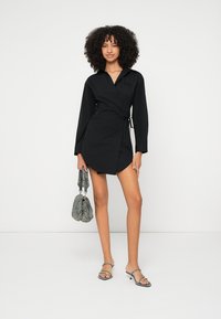 Čierne zavinovacie šaty s dlhými rukávmi, opaskom v páse a vysokým rozparkom. Doplnkové textúrovaná sivá kabelka a sandále na remienky.