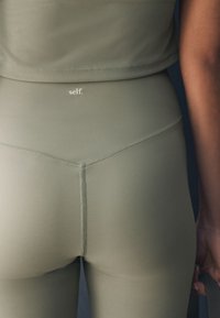 Oliiviroheline sportlik lühike topp ja kõrge vöökohaga retuusid, millel on sile tekstuur ning kaelal trükitud brändinimi „self.”
