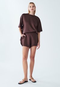 Brown cotton blend oversized t-shirt paired with matching shorts, featuring a side pocket and relaxed fit. Worn with black sandals.