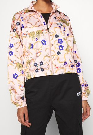 Femme portant une veste rose zippée avec une chaîne dorée et un imprimé léopard, des fleurs bleues et un pantalon noir avec un patch logo Adidas.