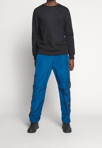 Felpa nera con collo rotondo, pantaloni sportivi blu lucidi con zip laterali e stivali neri. Design semplice, senza motivi o accenti prominenti.