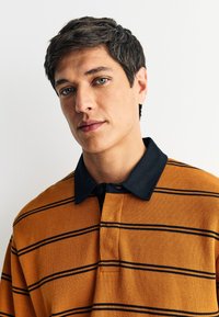 Prugasta polo majica u narančastoj i crnoj boji, s crnim ovratnikom, kopčanjem na gumbe i teksturiranim tkanjem. Prikaz uvećanog dijela oko vrata.