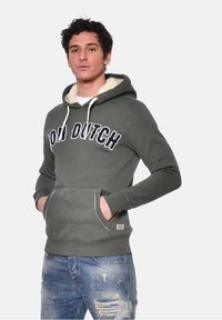 Sweatshirt à capuche vert olive avec cordons blancs, doublure en polaire, poche avant et graphique "HOLLAND" en bleu marine. Jean en denim déchiré.