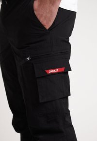 Černé cargo kalhoty s kapsou na zip a červenou nášivkou "JACKIT". Mají strukturovanou tkaninu a volný střih.