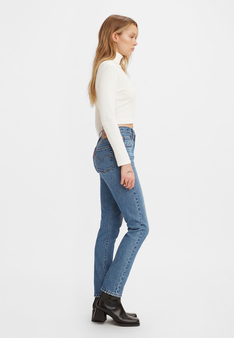 Schmale, dunkelblaue Jeans mit hoher Taille, kombiniert mit einem weißen langärmeligen Oberteil. Schwarze Absatz-Stiefeletten vervollständigen das Outfit.