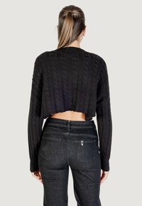 Donna con capelli castani lisci raccolti in una coda di cavallo, che indossa un maglione cropped a treccia nero e jeans neri a vita alta, in piedi di spalle.