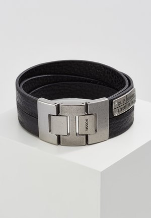 Bracelet en cuir noir texturé enroulé avec une large fermeture argentée mate gravée de "FOSSIL" et une petite plaque métallique avec des codes alphanumériques.