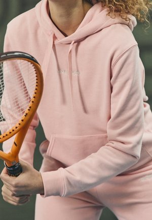 Personne portant un sweat à capuche rose tenant une raquette de tennis orange, se préparant à frapper un coup en intérieur.
