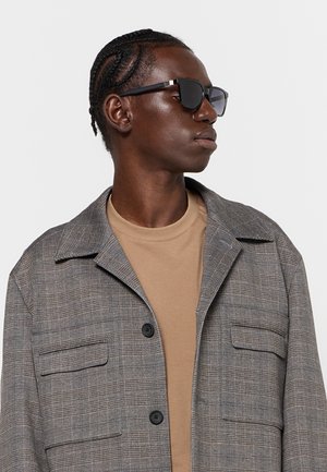 Uomo con i capelli intrecciati che indossa occhiali da sole neri, una camicia beige e una giacca grigia a quadri, rivolto di lato contro uno sfondo semplice.
