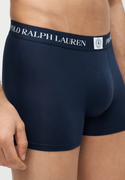 Prim-plan al unei persoane care poartă chiloți boxer Polo Ralph Lauren bleumarin, cu talie cu logo și o etichetă de marcă.