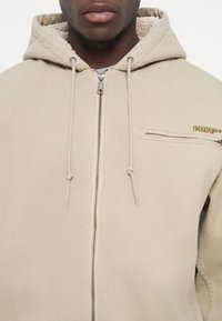 Beige hættejakke med lynlås foran, fleeceforing og en brystlomme med et lille guld logo. Snører på hætten er synlige.