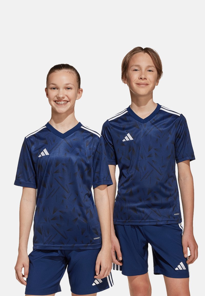 Due bambini che indossano maglie da calcio Adidas blu navy abbinate. Le maglie presentano un motivo geometrico nero e strisce bianche sulle spalle.