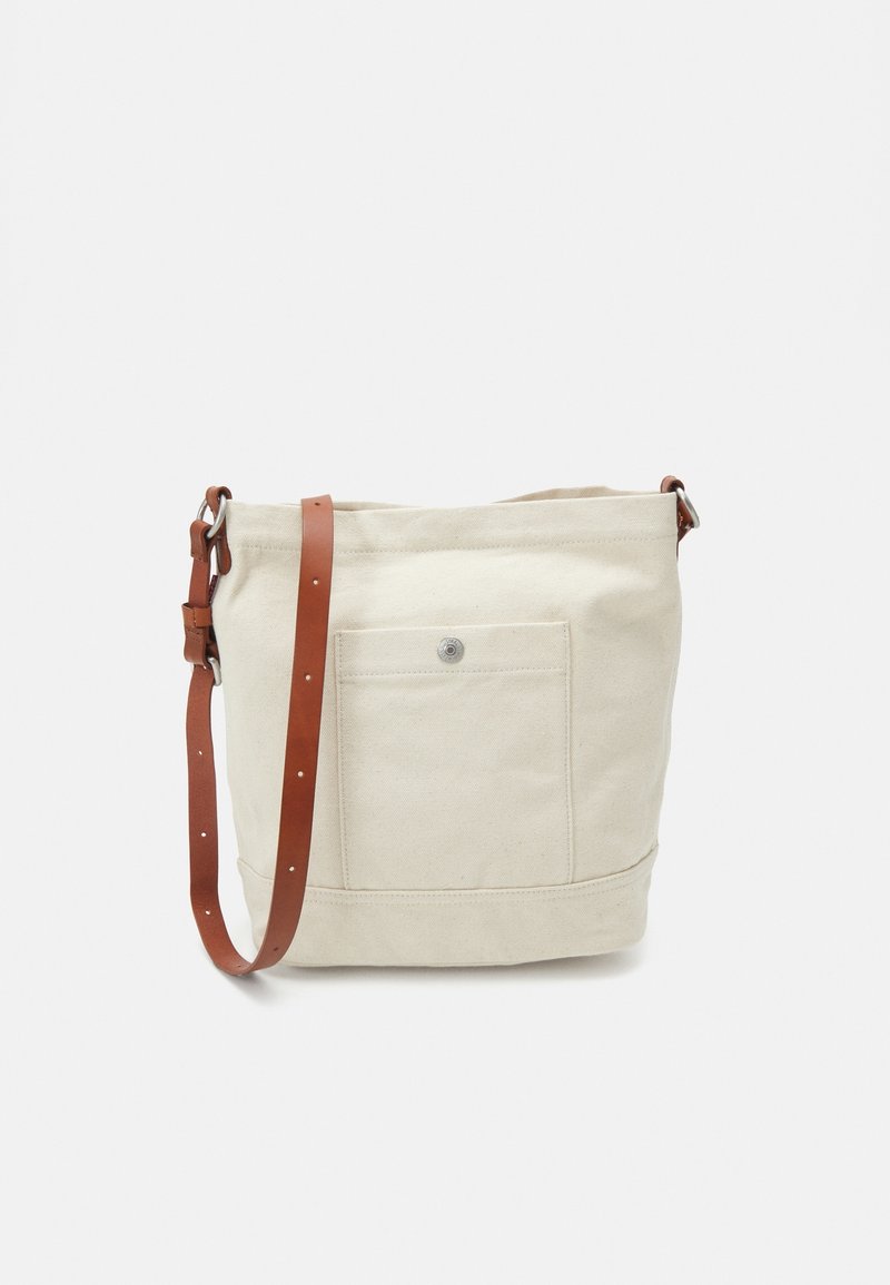 Beige Canvas-Eimerhandtasche mit Vordertasche und verstellbarem braunem Leder-Schulterriemen auf einfarbigem Hintergrund.