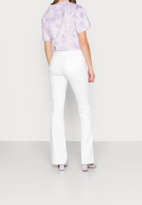 Valkoiset leveälahkeiset farkut yhdistettyinä liilan väriseen t-paitaan, jossa on solmuvärjäyskuvio, mikä luo rentoa ilmettä ja sileää tekstuuria. Takana on taskut ja vaatteessa on hieman kiiltoa.