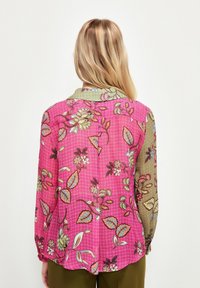 Blouse à manches longues avec un motif floral rose, un col vert, et un tissu léger et texturé. Présente un ourlet incurvé et des accents floraux.