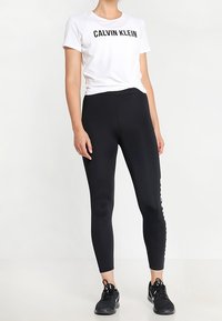 Mujer vestida con una camiseta blanca de Calvin Klein, leggings negros de talle alto con logo lateral y zapatillas deportivas negras, de pie contra un fondo blanco.