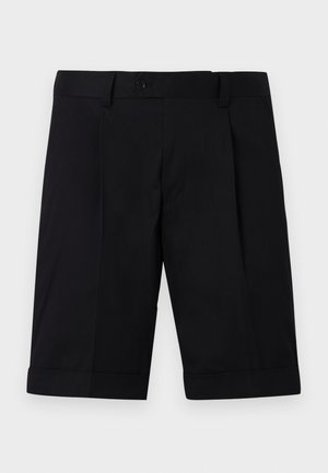 Man wearing olive green short-sleeve zip-collar shirt, black knee-length shorts, white socks, and beige sneakers standing with hands in pockets.