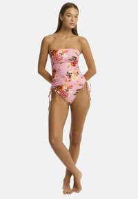 Un modèle se tient avec confiance dans un maillot de bain floral rose sans bretelles, avec des liens sur les côtés, mettant en valeur un look estival sur un fond blanc.