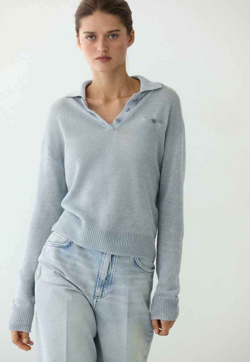 Giovane donna con capelli raccolti che indossa un maglione a maglia grigio chiaro con collo e jeans a vita alta azzurri, in piedi davanti a uno sfondo uniforme.