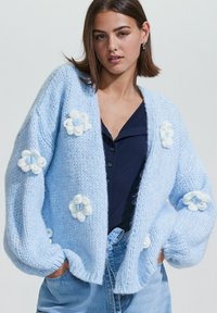 Un mannequin pose en portant un cardigan oversize bleu clair orné de fleurs tricotées blanches, associé à une chemise sombre et un jean en denim. Fond neutre.