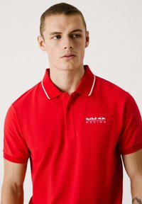 Crvena polo majica s ovratnikom, s bijelim detaljima. Logotip Red Bull Racing vidljivo prikazan na lijevoj strani prsnog područja. Glatka tekstura tkanine.