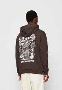 Sweat à capuche marron avec un grand imprimé graphique blanc au dos, représentant des fleurs, un visage souriant, et le texte "CONTINUE D'AIDER À GRANDIR."