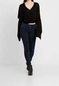 Haut noir à manches longues avec col en V et taille cintrée, associé à un jean skinny bleu foncé et des bottines noires. Design simple et ajusté.