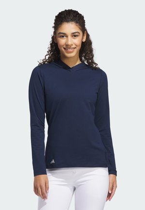 Jonge vrouw met krullend haar die een donkerblauw capuchontrui met lange mouwen en een witte broek draagt, glimlachend en staand voor een effen achtergrond.