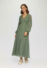 Robe longue verte en tissu léger, avec un décolleté en V, des manches bouffantes et un ourlet à volants. Assortie à des chaussures beiges épaisses.