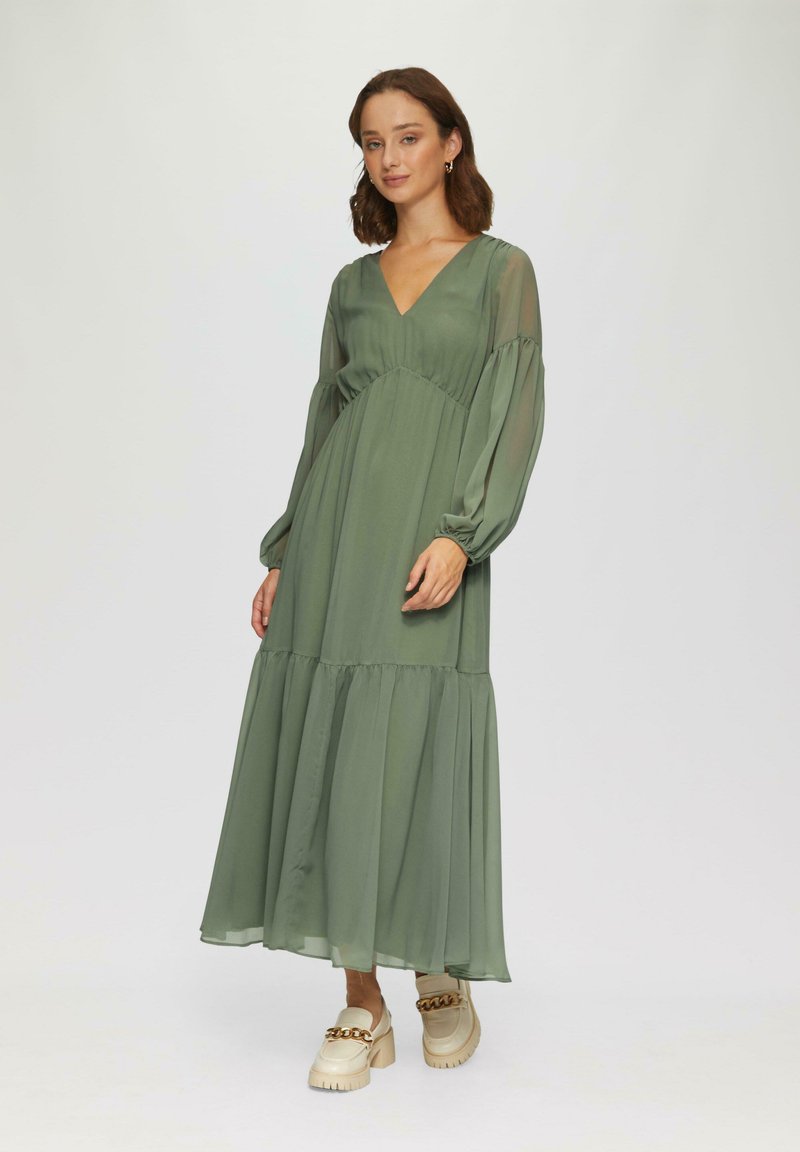 Robe longue verte en tissu léger, avec un décolleté en V, des manches bouffantes et un ourlet à volants. Assortie à des chaussures beiges épaisses.
