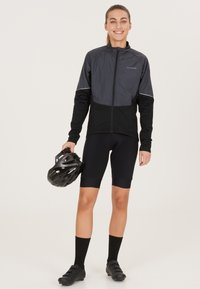 Svart cykeljacka med grå detaljer, framzipper och långa ärmar. Bärs med svarta shorts och svarta cykelskor. Håller en svart hjälm.