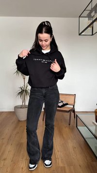 Noor naine siseruumis reguleerib musta Tommy Hilfigeri kapuutsi, kannab tumedaid teksaseid ja tenniseid, seisab puidust põrandal taimede ja mööbli lähedal.