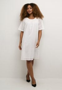 Rochie din bumbac alb, cu broderie din ochiuri, decolteu prins și mâneci scurte în formă de clopot. Culoarea este mai sus de genunchi. Pantofi cu toc negri completează ținuta.