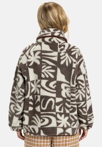 Veste en polaire marron et crème avec un motif géométrique et floral. Col montant et coupe légèrement oversize, présentant un tissu texturé.