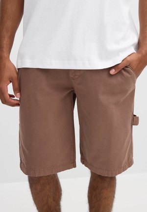 Hombre vistiendo pantalones cortos marrones hasta la rodilla y una camiseta blanca, con las manos cerca de los bolsillos, de pie contra un fondo claro y sencillo.