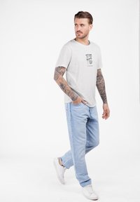 Hombre con tatuajes en las mangas completo caminando, vistiendo una camiseta gráfica blanca, jeans celestes y zapatillas blancas, mirando hacia su izquierda.