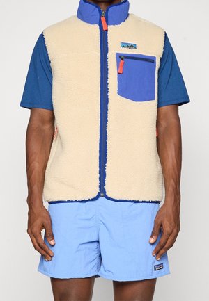 Mand iført beige fleecevest med blå lynlås og brystlomme over blå t-shirt og lyseblå shorts.